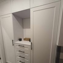 Biała zabudowa meblowa z ciemnymi uchwytami, obejmująca szafę z przesuwnymi drzwiami i komodę z szufladami, uzupełniona otwartą półką z drobnymi przedmiotami.