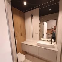 Nowoczesna łazienka z zabudową meblową z jasnego drewna, białą umywalką nablatową o owalnym kształcie i minimalistyczną armaturą w kolorze czarnym; w lustrze odbija się osoba robiąca zdjęcie.