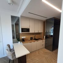 Niewielka kuchnia w jasnych barwach, z blatem drewnopodobnym, czarną armaturą i zabudową sięgającą sufitu. Widoczna mała jadalnia z blatem i krzesłem. Podłoga w jodełkę.