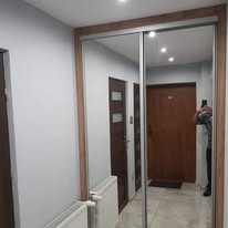 Szafa wnękowa z lustrzanymi drzwiami przesuwnymi w przedpokoju, widoczne grzejniki i drzwi wejściowe, osoba robiąca zdjęcie w odbiciu.