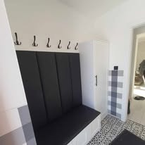 Wnętrze przedpokoju w Dąbrowie Górniczej z siedziskiem tapicerowanym na czarno, wieszakami na ścianie i białą szafką. Na ścianie widoczny wzór w kratę.