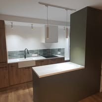 Nowoczesna kuchnia z drewnianymi szafkami, białym zlewem farmerskim i zielonymi płytkami nad blatem. Wyspa kuchenna w kolorze oliwkowym z białym blatem.