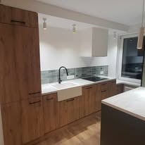 Nowoczesna kuchnia z drewnianymi frontami szafek, białym zlewem farmerskim i szarymi płytkami nad blatem. Widoczne minimalistyczne oświetlenie punktowe i biały okap. Blat jasny, imitujący kamień.