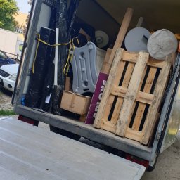 ERYK KOC TRANSPORT - Otwarta paka busa wypełniona różnorodnymi przedmiotami: paleta, opakowane elementy, karton, okrągłe podstawy, elementy ekspozycyjne. Widoczne drzewa i budynki w tle.