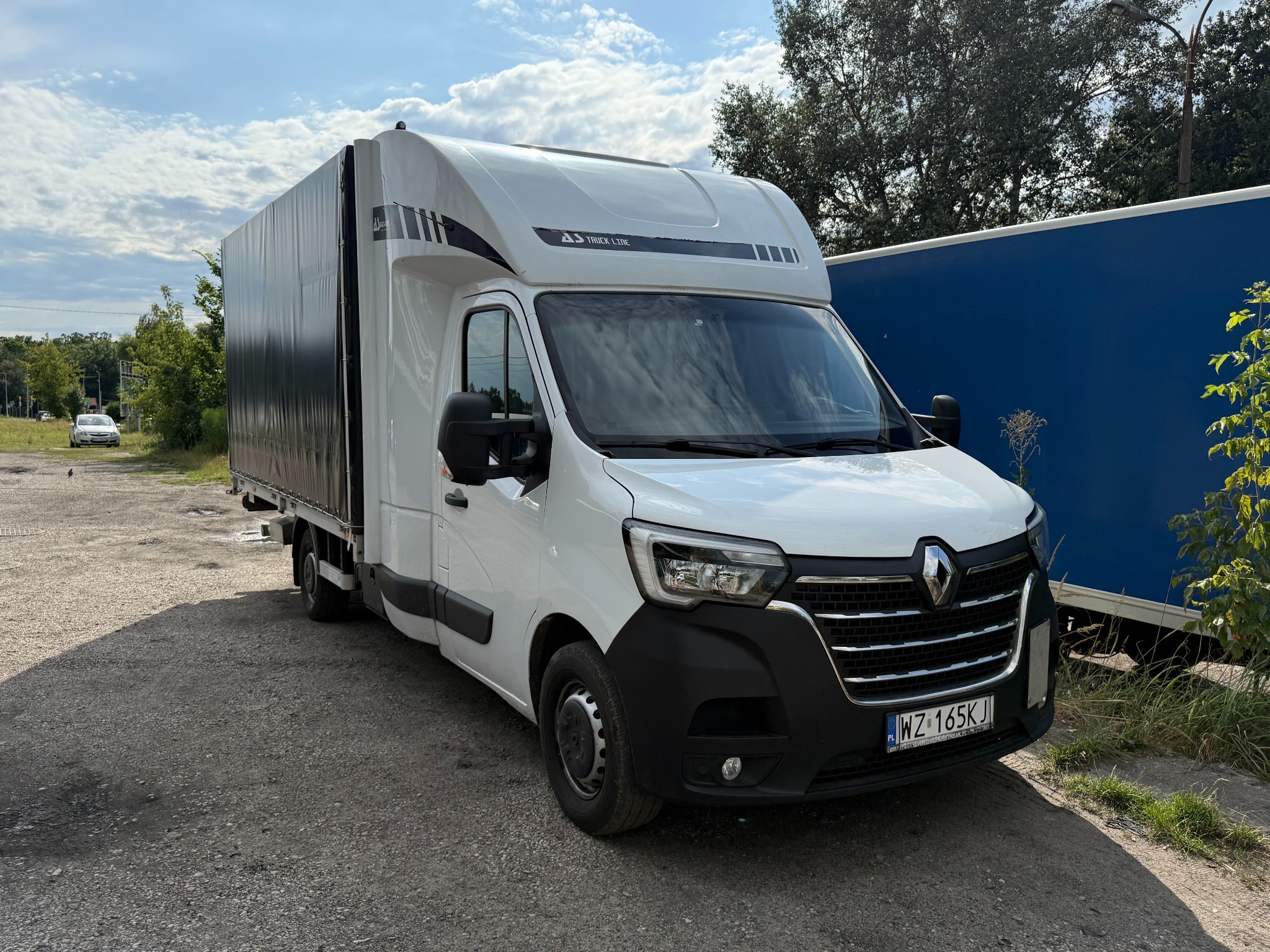 Biały bus Renault Master z plandeką na parkingu, widok z przodu, tablica rejestracyjna WZ 165KJ, niebieska naczepa w tle, Truck Line.