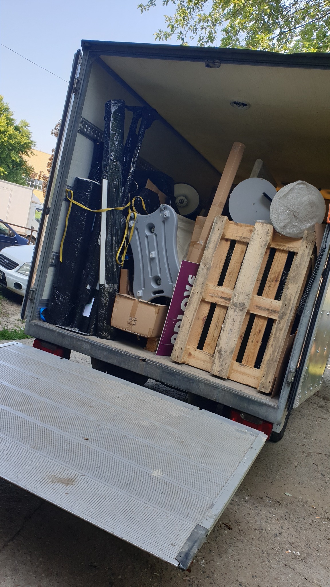 Otwarta paka busa wypełniona różnorodnymi przedmiotami: paleta, opakowane elementy, karton, okrągłe podstawy, elementy ekspozycyjne. Widoczne drzewa i budynki w tle.