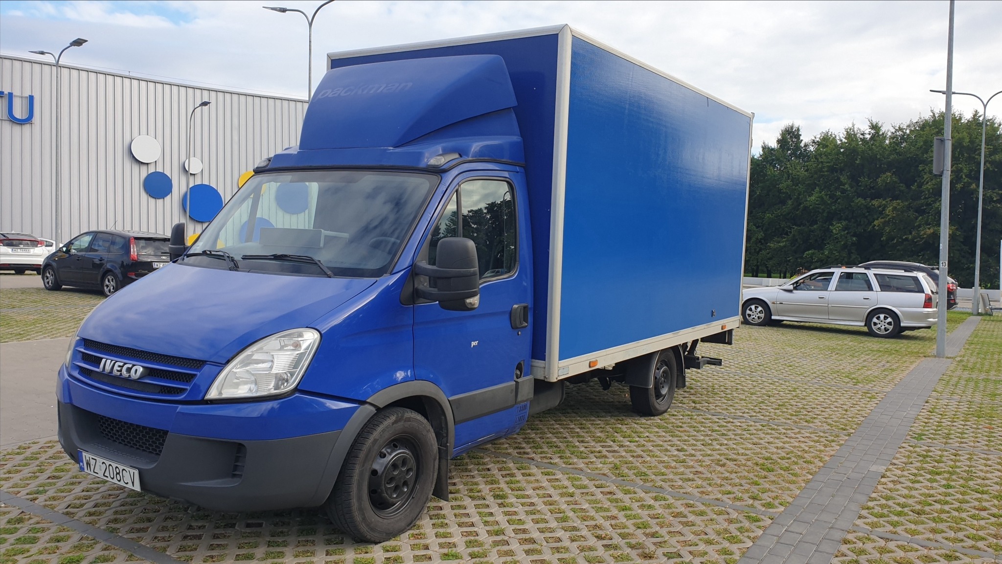 Niebieski samochód dostawczy marki Iveco z nadrukiem 'packman' na parkingu z kostki betonowej, na tle budynku z logo 'U' i zieleni.