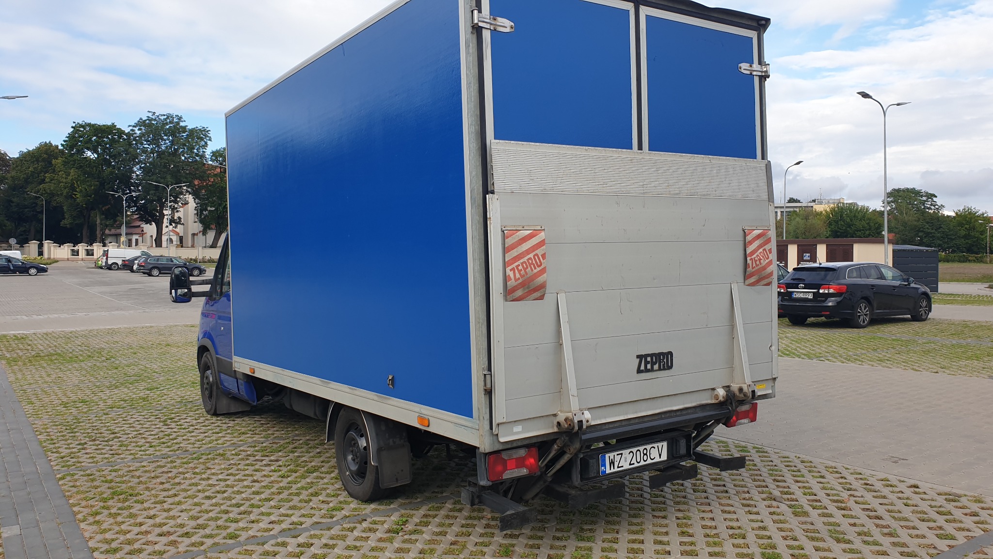 Niebieski samochód dostawczy z podnośnikiem ZEPRO na parkingu z ażurowej kostki, widok z tyłu.
