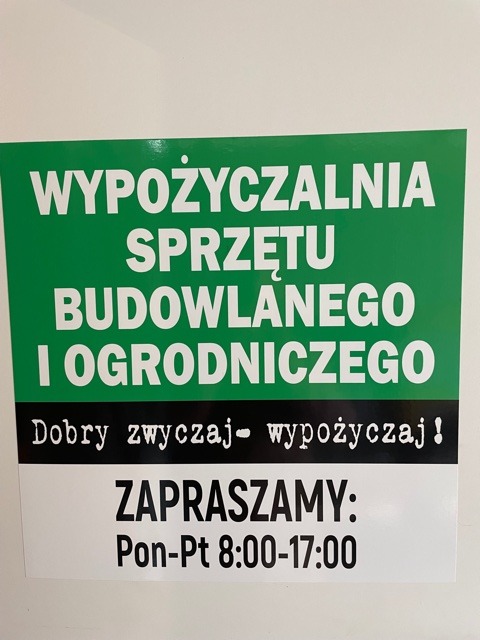 Zielono-biały szyld informacyjny z napisem 'Wypożyczalnia sprzętu budowlanego i ogrodniczego', hasłem 'Dobry zwyczaj - wypożyczaj!' oraz godzinami otwarcia: Pon-Pt 8:00-17:00.