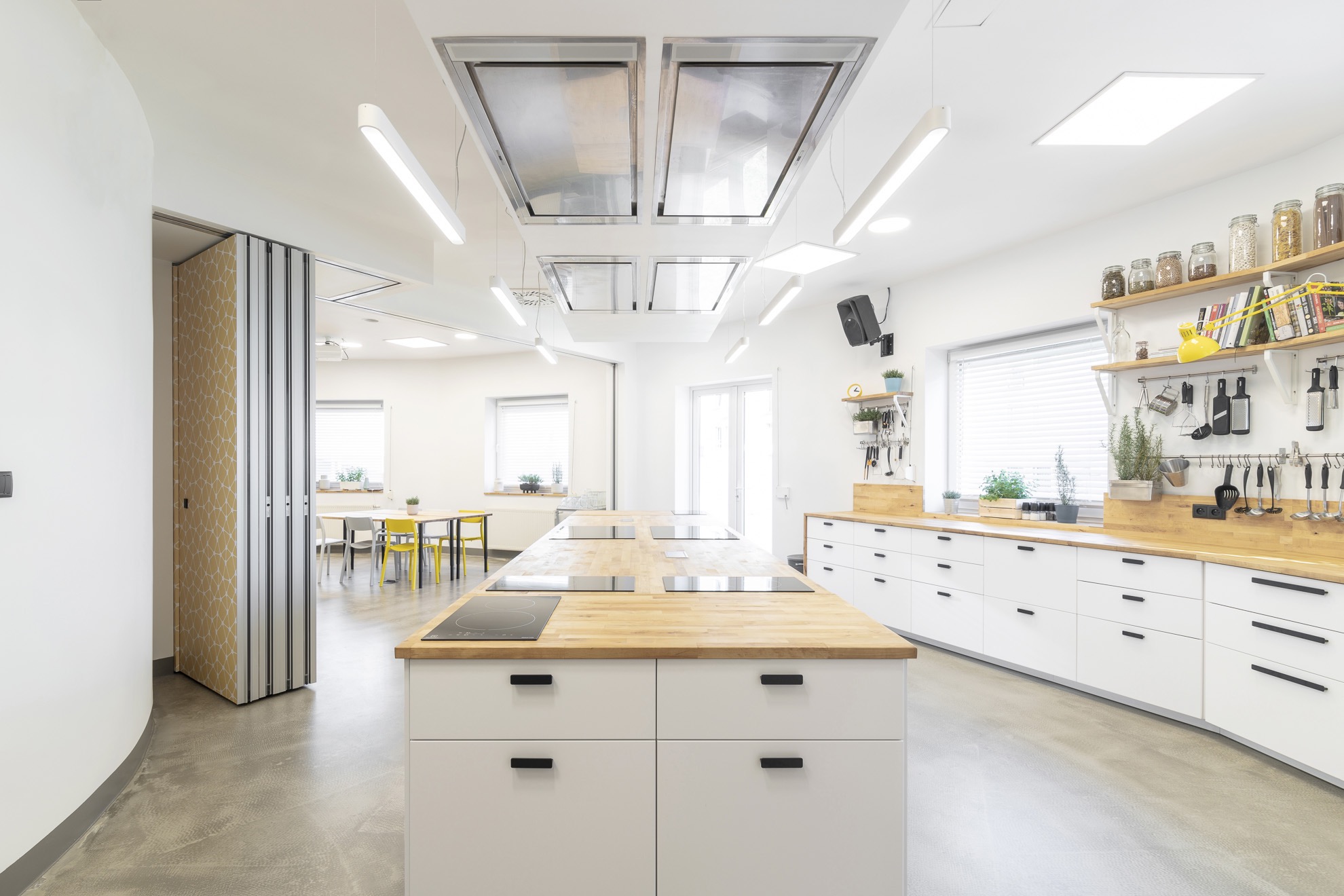 Jasna kuchnia z wyspą i płytami indukcyjnymi, drewnianymi blatami, białymi szafkami i otwartą przestrzenią z żółtymi krzesłami.