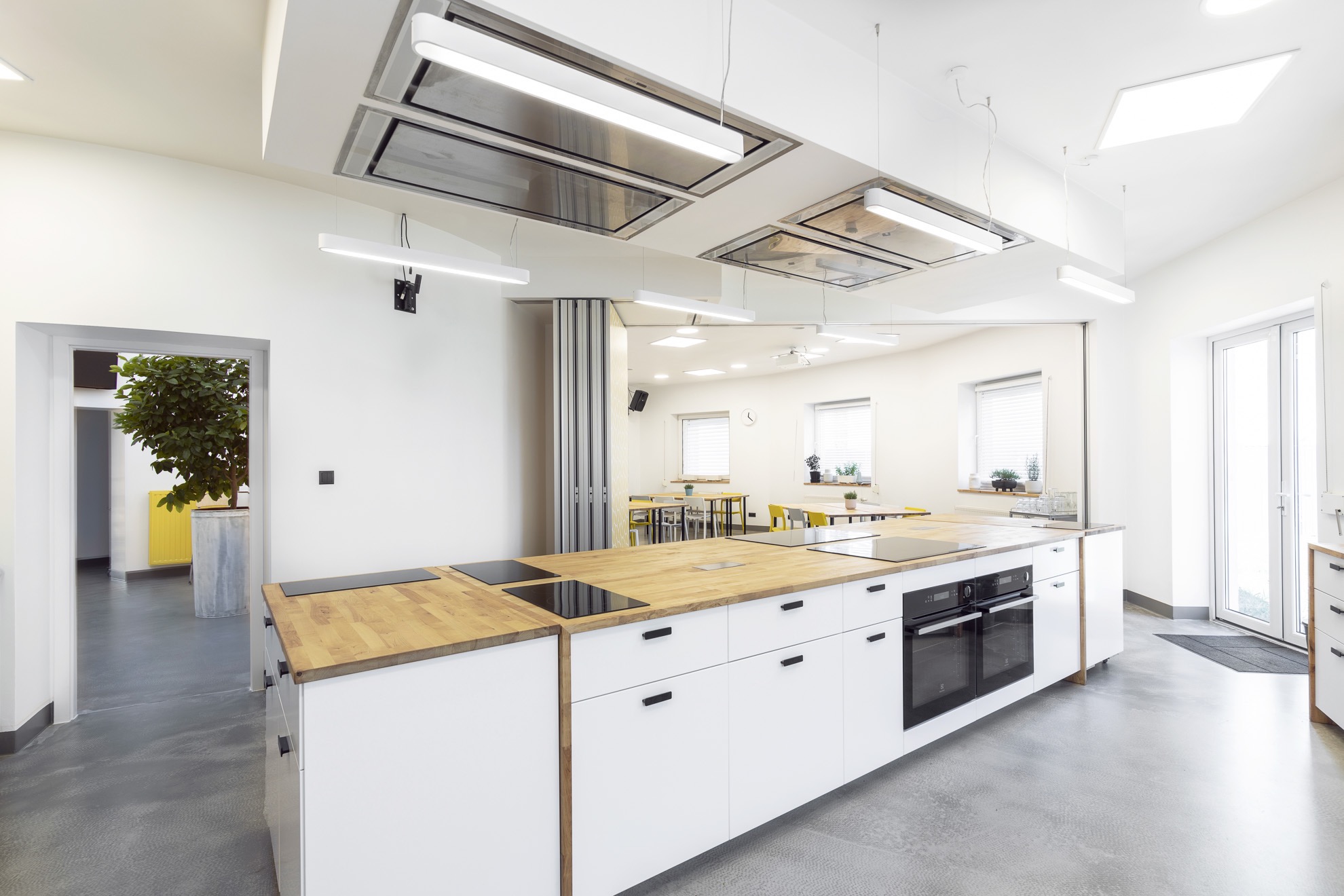 Jasne, przestronne wnętrze z dużą wyspą kuchenną z drewnianym blatem i białymi szafkami, widok na salę konferencyjną z żółtymi krzesłami w tle.