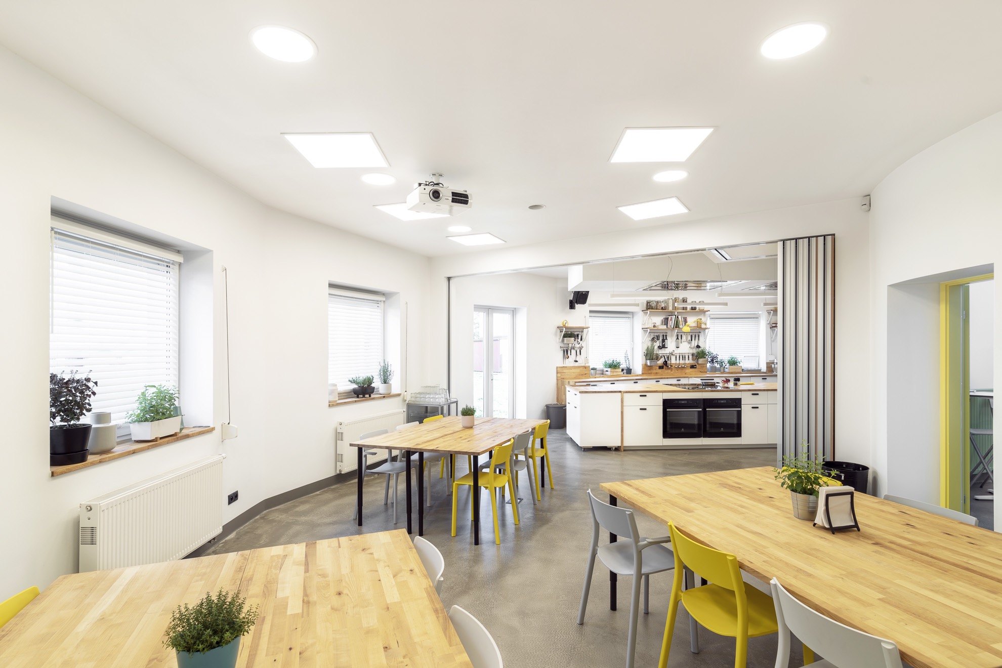 Jasne, przestronne wnętrze z drewnianymi stołami, żółtymi i szarymi krzesłami, otwartą kuchnią w tle i projektorem zamontowanym na suficie.