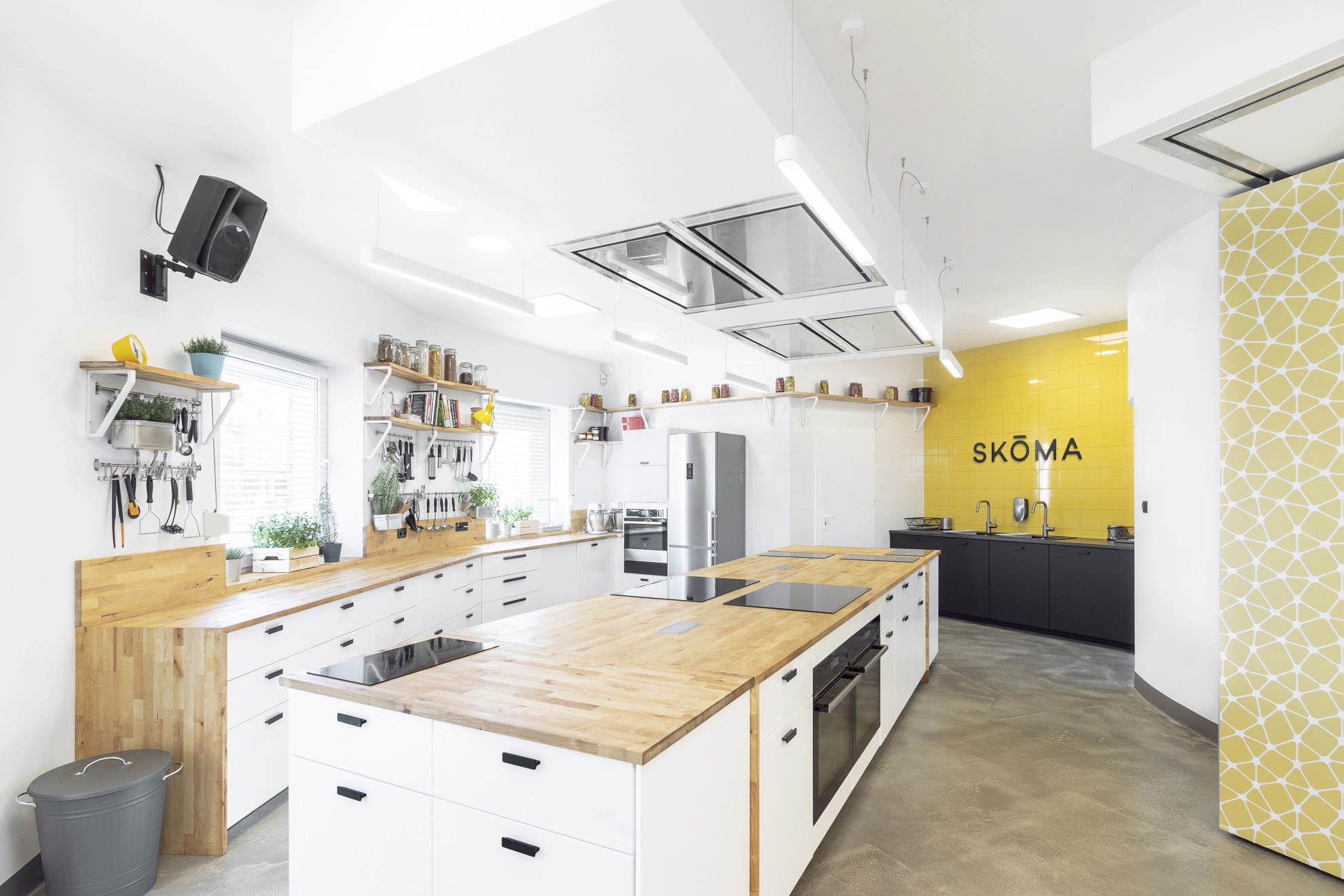 Jasna, nowoczesna kuchnia z wyspą i drewnianymi blatami, żółta ściana z napisem SKÖMA, głośnik na ścianie po lewej stronie.