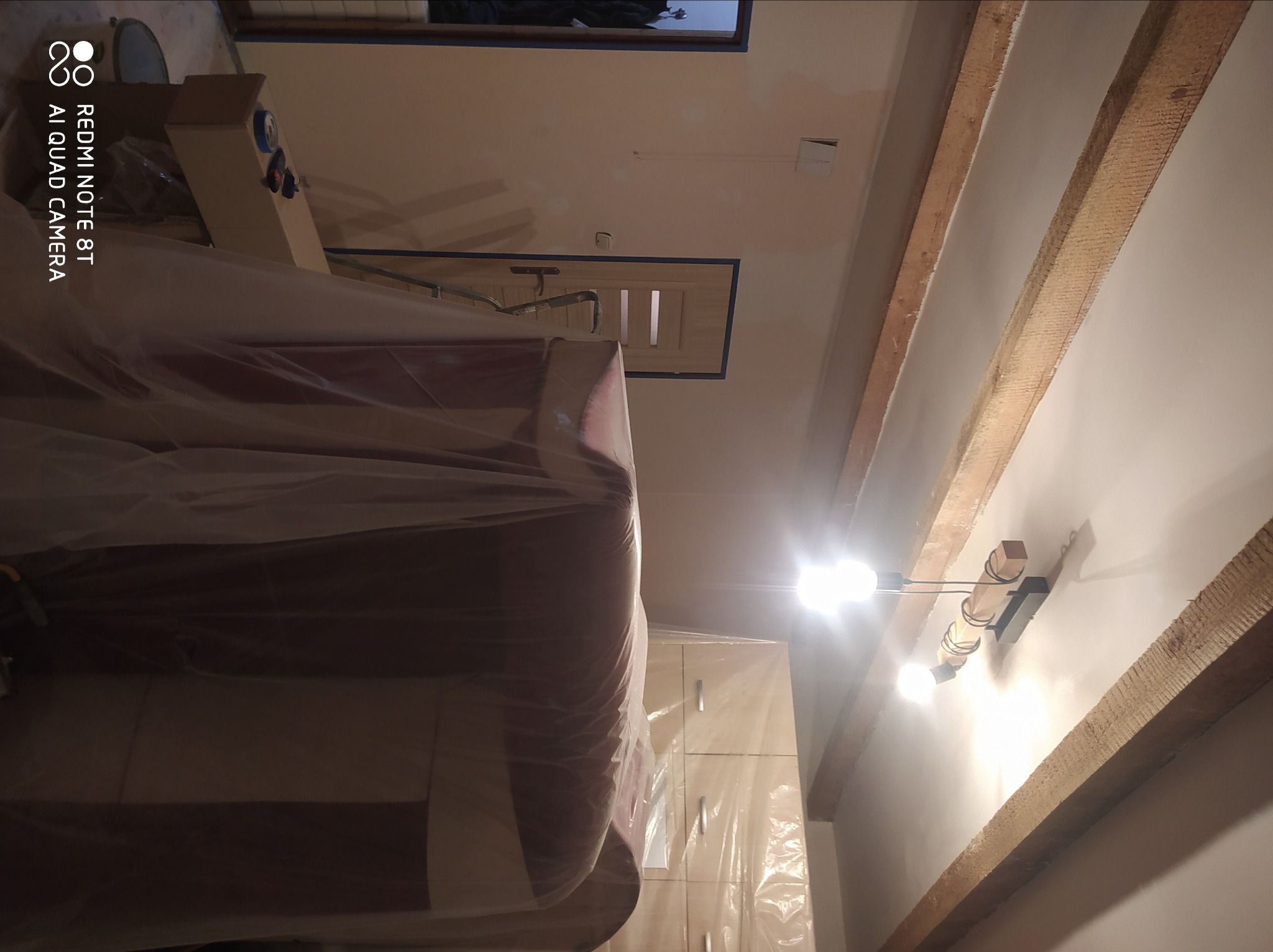 Pokryty folią mebel w trakcie remontu pomieszczenia z drewnianymi belkami stropowymi i nową lampą sufitową.