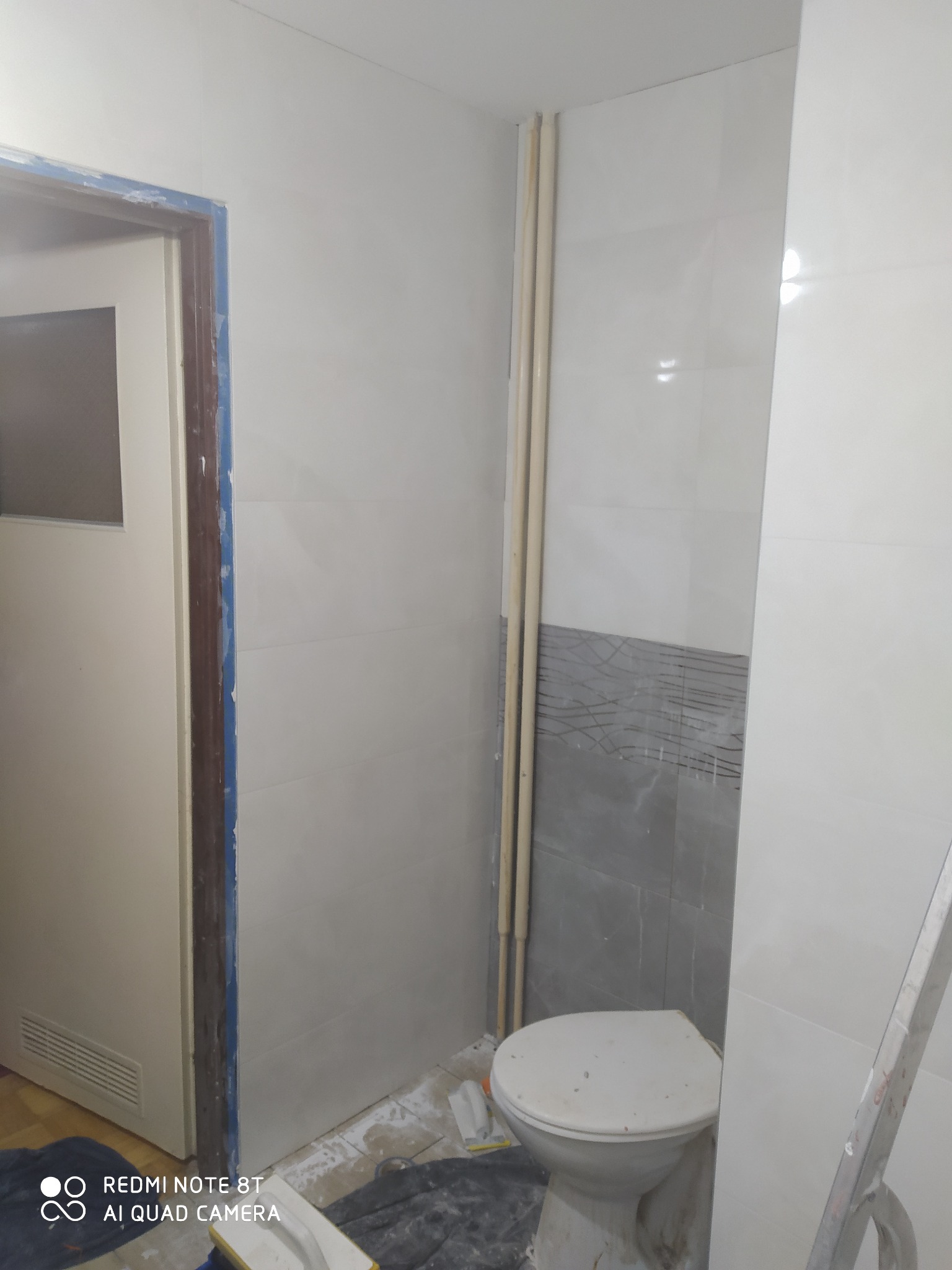Wnętrze łazienki w trakcie remontu: białe płytki na ścianach z szarym pasem dekoracyjnym, widoczne rury pionowe, toaleta oraz futryna drzwi zabezpieczona taśmą malarską, na podłodze widoczne...