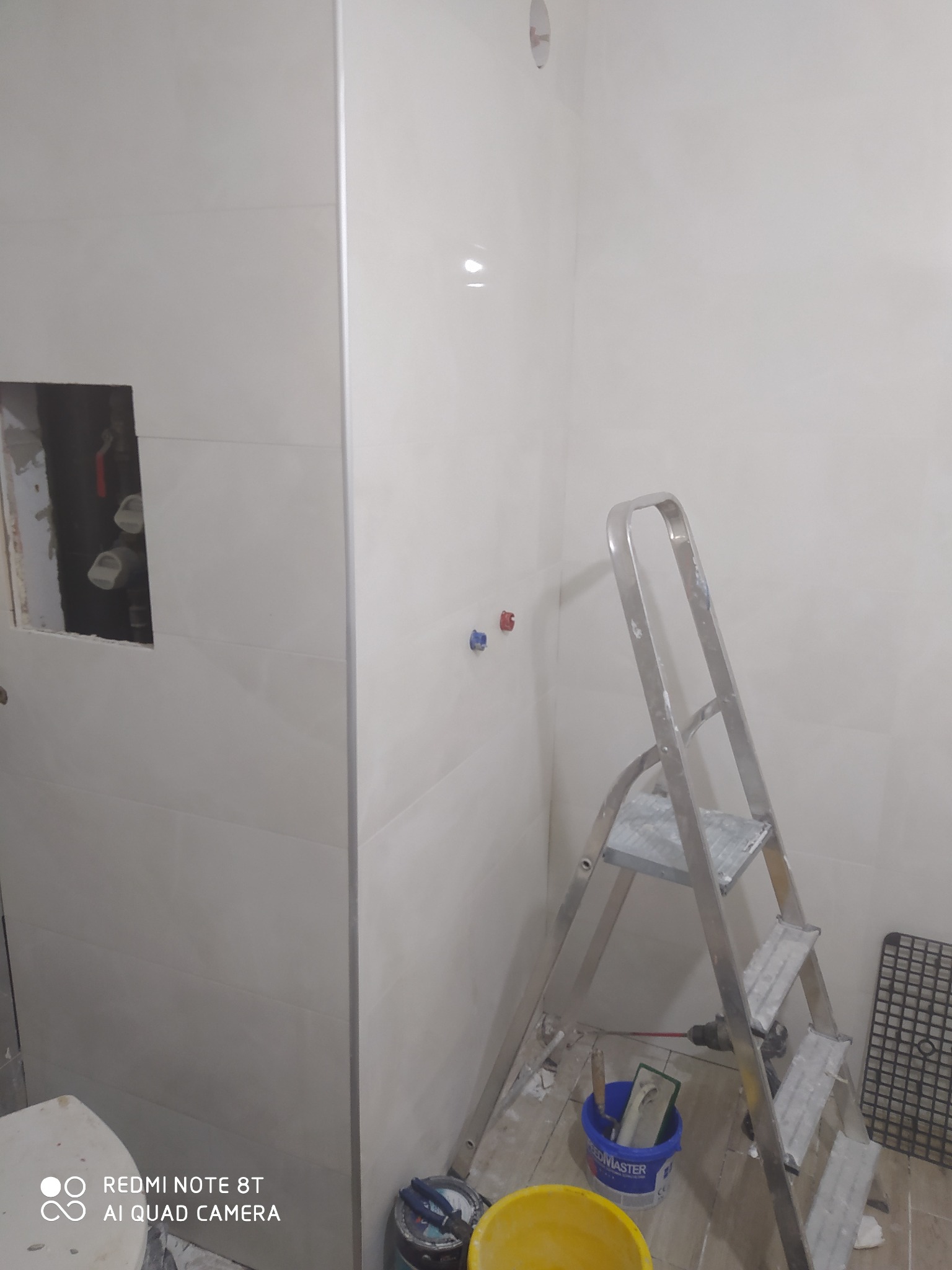 Wnętrze łazienki w trakcie remontu, z widocznymi jasnymi, prostokątnymi płytkami na ścianach, otworem na instalację, aluminiową drabiną, wiadrami i narzędziami na podłodze.