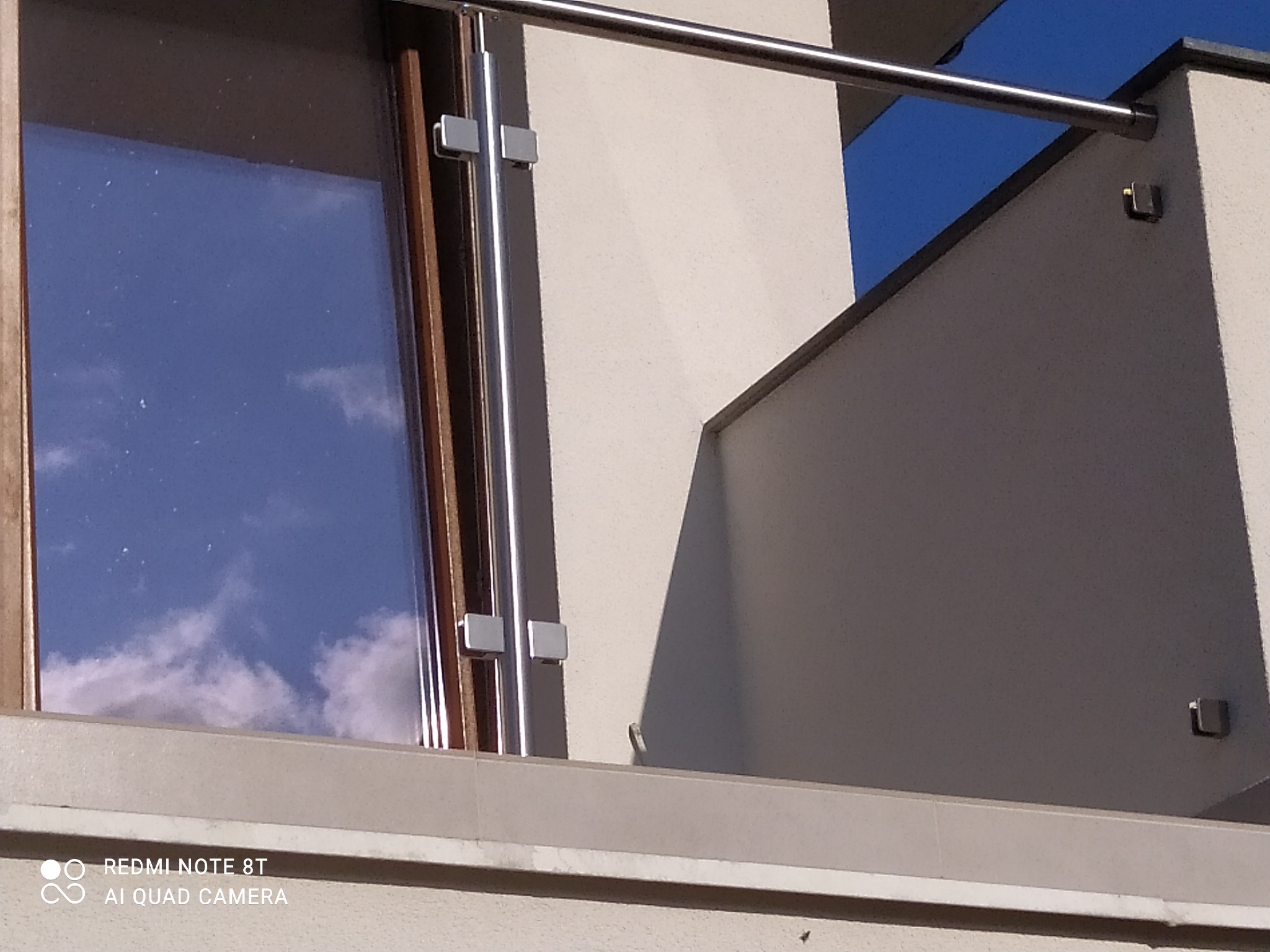Fragment balkonu z nowoczesną balustradą ze szkła i stali nierdzewnej, mocowaną do ściany budynku; w tle błękitne niebo z odbijającymi się chmurami w szybie okiennej.