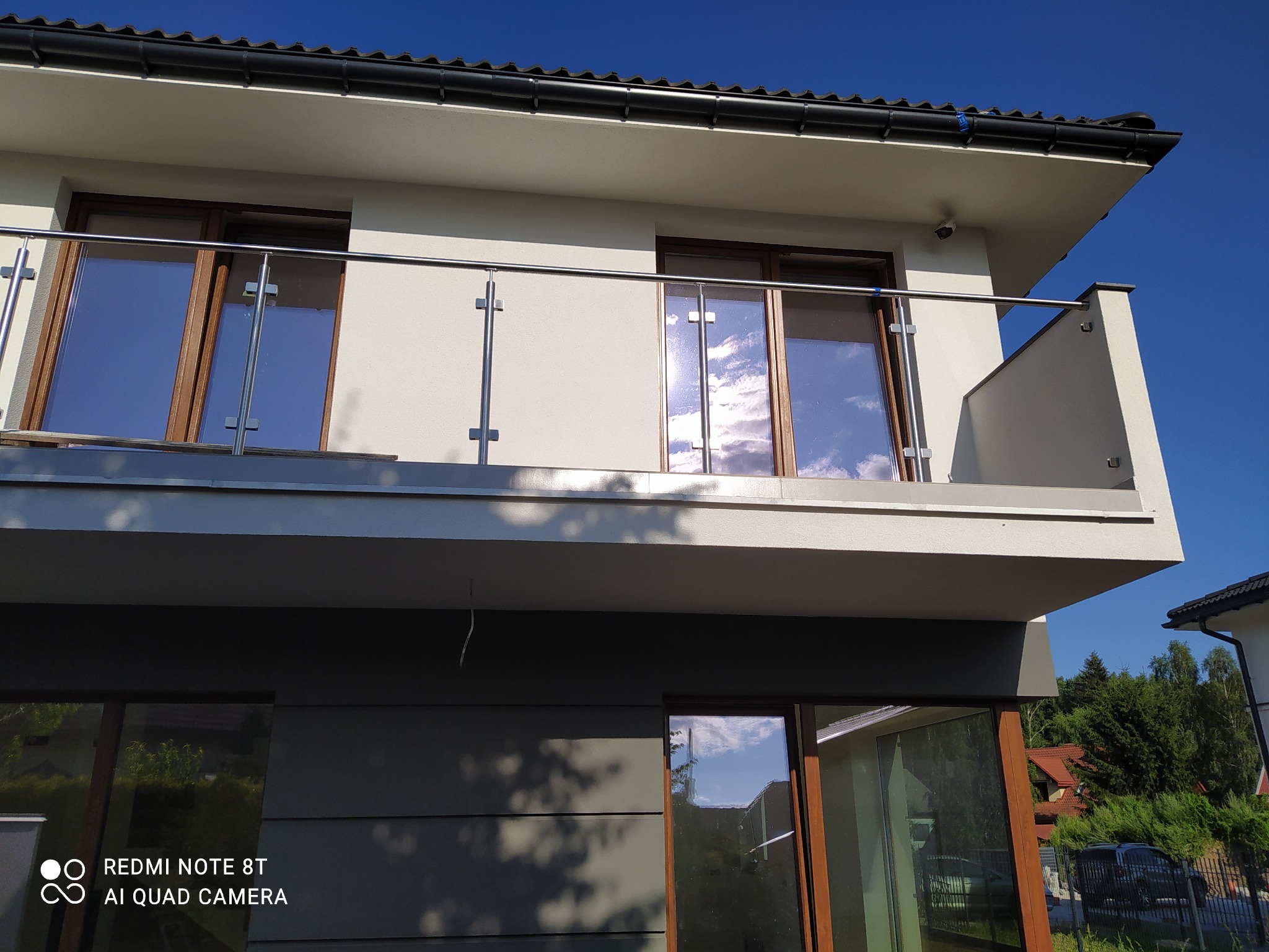 Nowoczesny balkon z balustradą ze stali nierdzewnej i szkła, odbijający błękitne niebo, na tle jasnej elewacji domu.