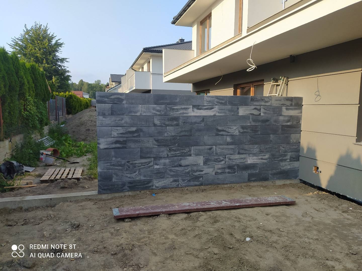 Wznoszona ściana z szarej, łupanej kostki betonowej na posesji, widoczne niedokończone prace ziemne i fragment elewacji domu.