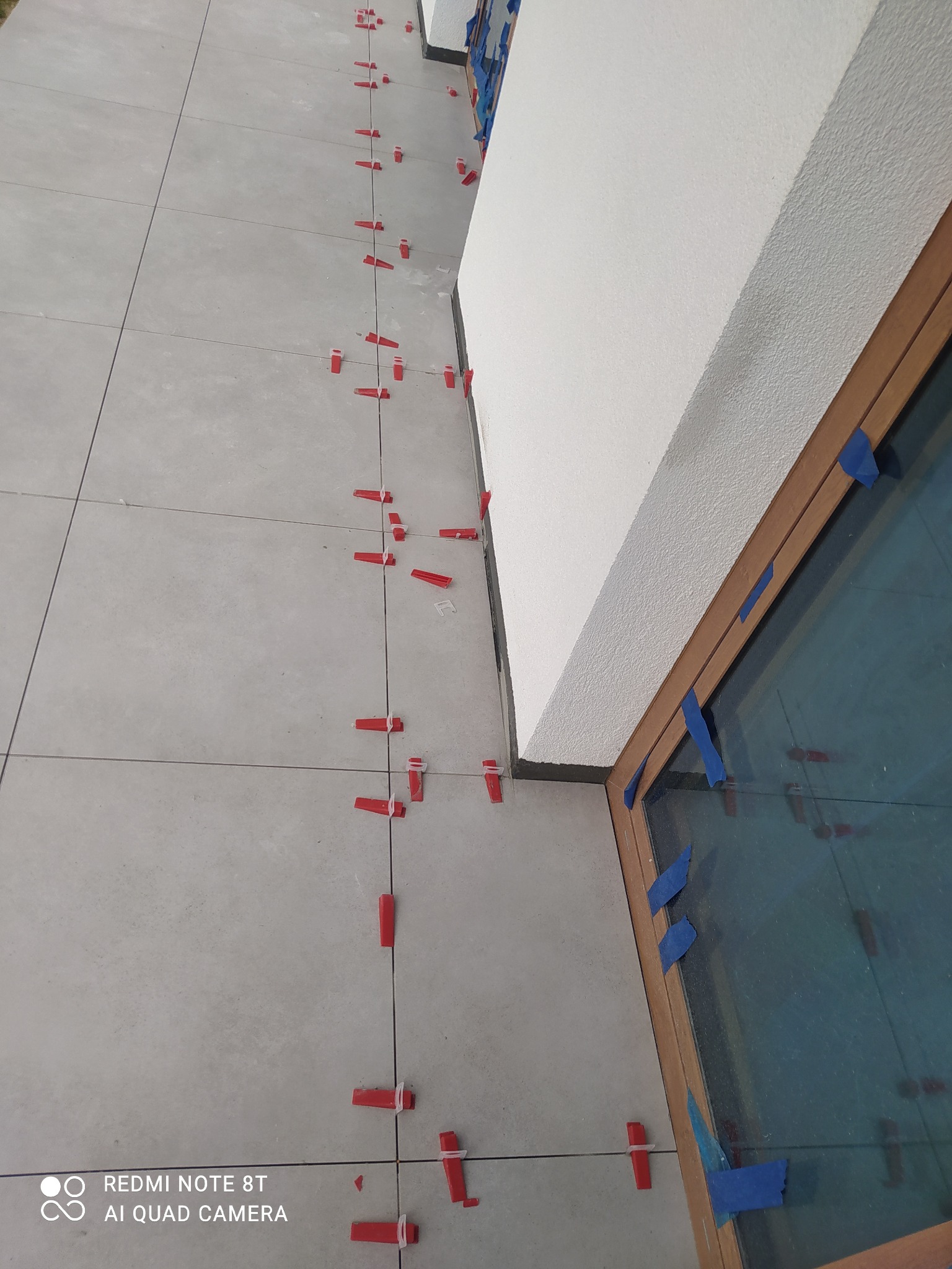 Szare płytki podłogowe z czerwonymi klipsami poziomującymi w trakcie montażu, widoczne niebieskie taśmy malarskie zabezpieczające ramę okienną