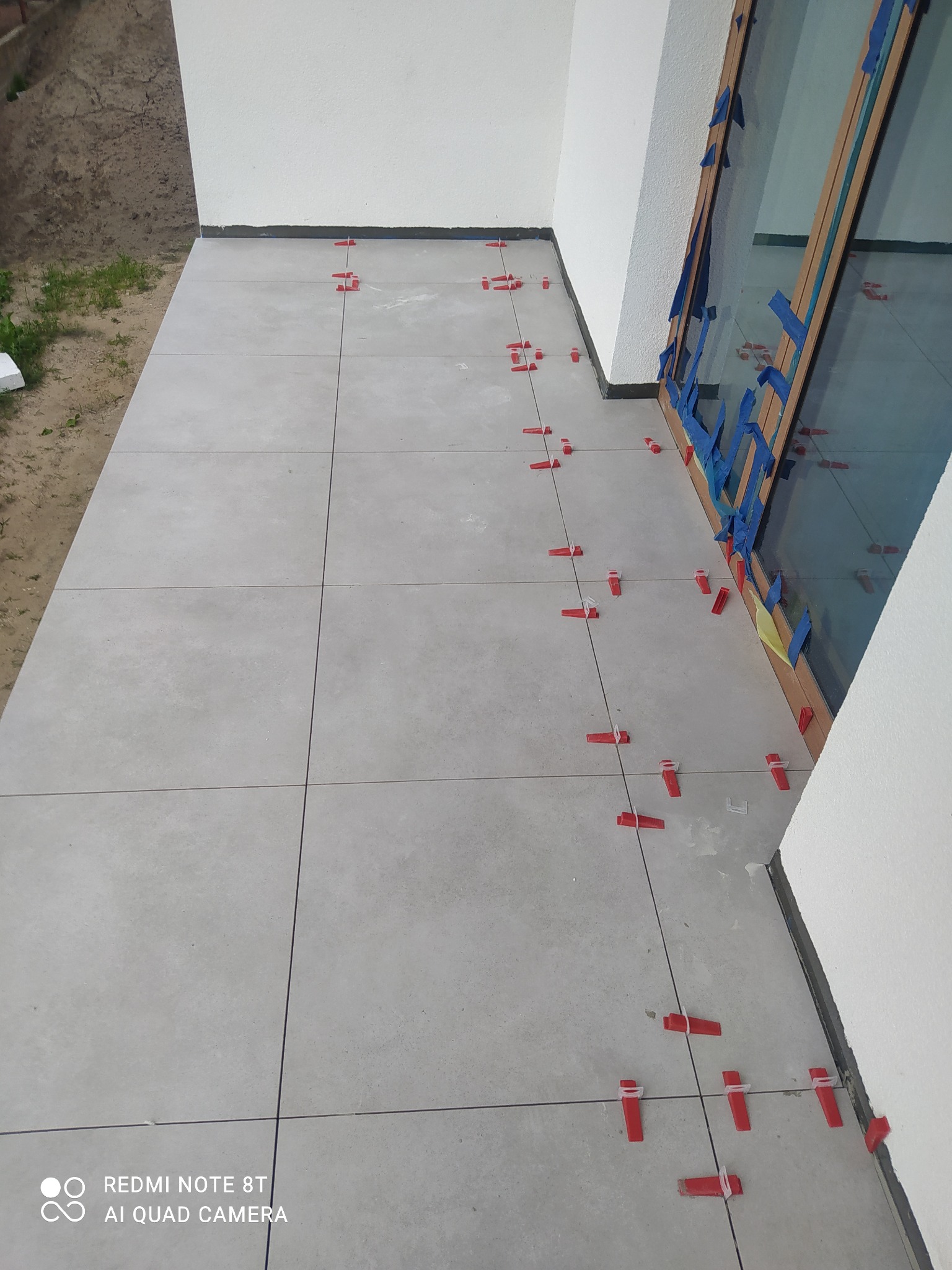 Jasnoszare płytki tarasowe ułożone na zewnątrz budynku, z czerwonymi klipsami poziomującymi widocznymi w spoinach, okno zabezpieczone niebieską taśmą malarską.