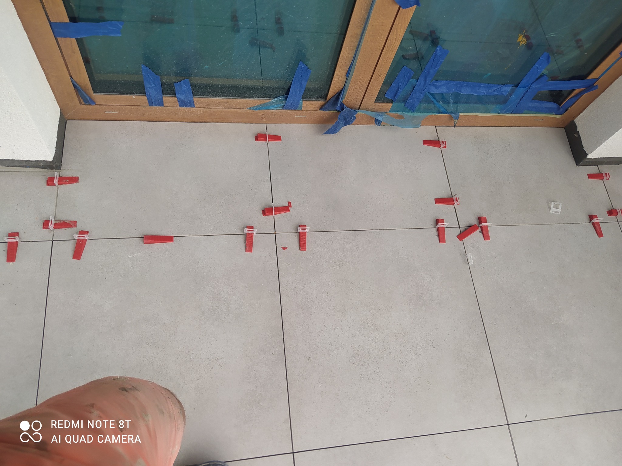 Jasne płytki podłogowe w trakcie układania, z widocznymi czerwonymi klipsami poziomującymi i sznurkiem wyznaczającym linie, przy oknie zabezpieczonym niebieską folią malarską.