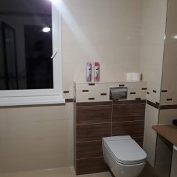 Michał Kukawa - Nowoczesna łazienka z podwieszaną toaletą, beżowymi i brązowymi płytkami, oraz oknem z białymi ramami. Na półce nad toaletą widoczne dwa odświeżacze powietrza i zapas papieru toaletowego.