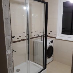 Michał Kukawa - Montaż kabiny prysznicowej z czarnymi profilami w łazience, widoczna pralka i fragment surowej ściany przed wykończeniem.