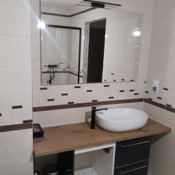 Michał Kukawa - Nowoczesna łazienka z umywalką nablatową, czarną armaturą i dużym lustrem z oświetleniem, płytki w odcieniach beżu z ciemnobrązowymi akcentami, widoczny fragment toalety.