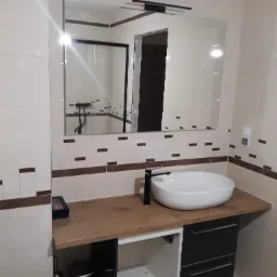 Nowoczesna łazienka z umywalką nablatową, czarną armaturą i dużym lustrem z oświetleniem, płytki w odcieniach beżu z ciemnobrązowymi akcentami, widoczny fragment toalety.