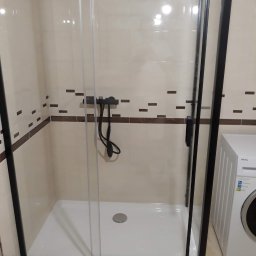 Michał Kukawa - Kabina prysznicowa z czarnymi profilami i przesuwnymi drzwiami, z widoczną armaturą i fragmentem pralki Amica w łazience wykończonej beżowymi płytkami z brązowymi akcentami.