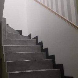 Michał Kukawa - Schody z szarych płytek, wykończone listwą, na ścianie tapeta w szaro-białe pasy oraz wzór geometryczny w romby.