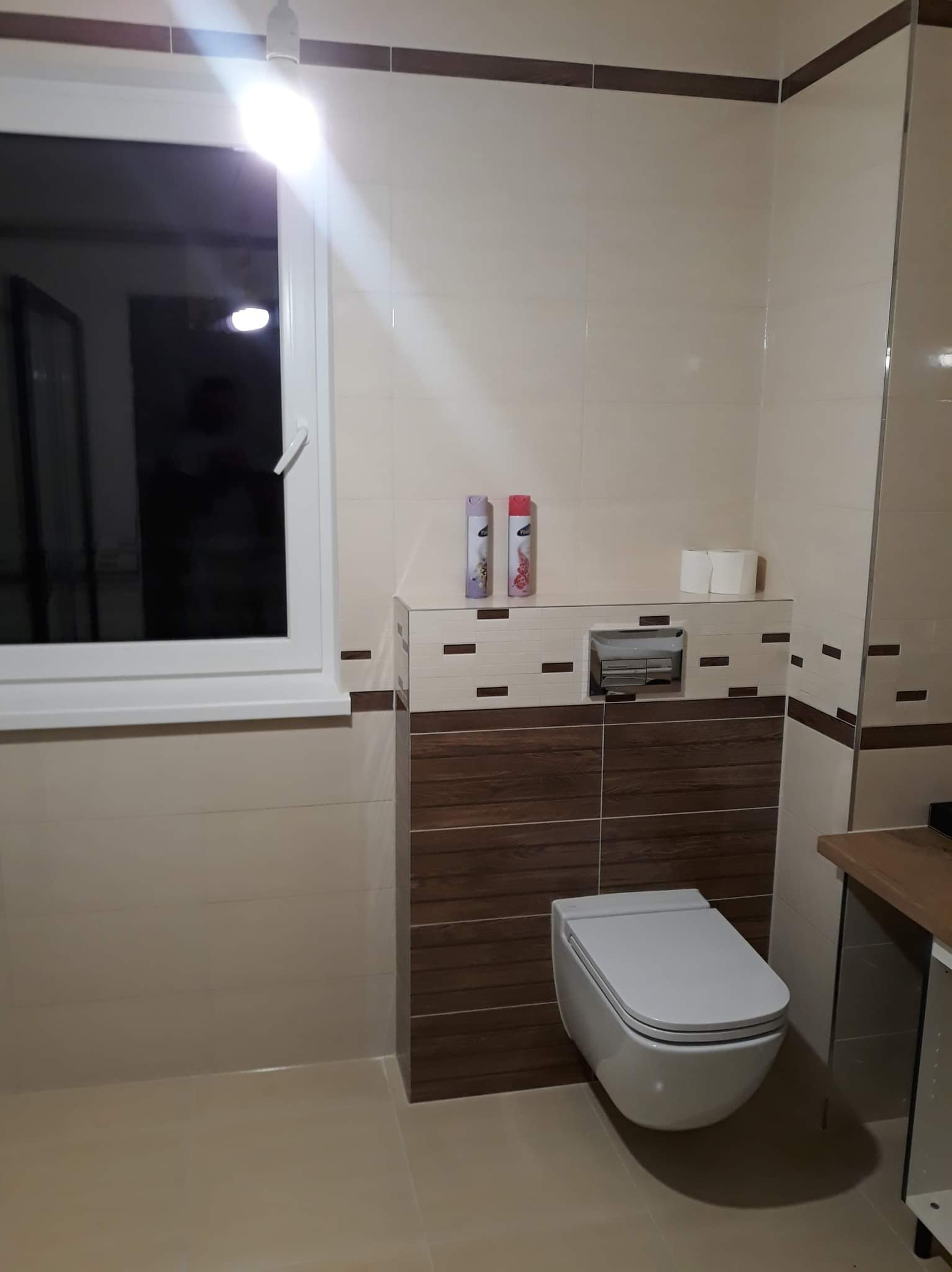 Nowoczesna łazienka z podwieszaną toaletą, beżowymi i brązowymi płytkami, oraz oknem z białymi ramami. Na półce nad toaletą widoczne dwa odświeżacze powietrza i zapas papieru toaletowego.