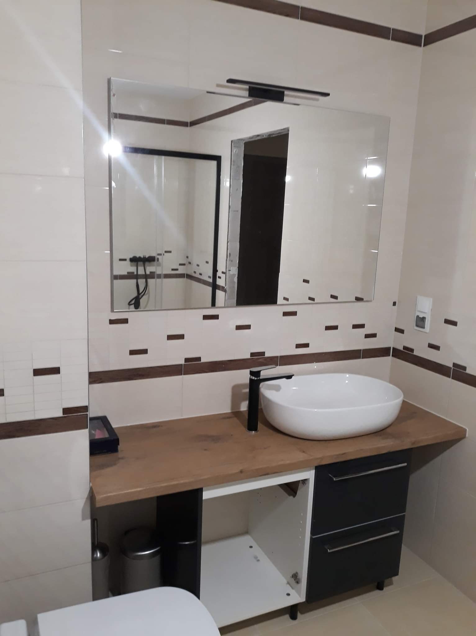 Nowoczesna łazienka z umywalką nablatową, czarną armaturą i dużym lustrem z oświetleniem, płytki w odcieniach beżu z ciemnobrązowymi akcentami, widoczny fragment toalety.