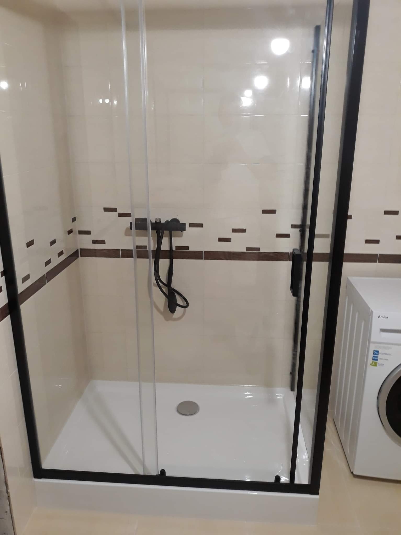 Kabina prysznicowa z czarnymi profilami i przesuwnymi drzwiami, z widoczną armaturą i fragmentem pralki Amica w łazience wykończonej beżowymi płytkami z brązowymi akcentami.