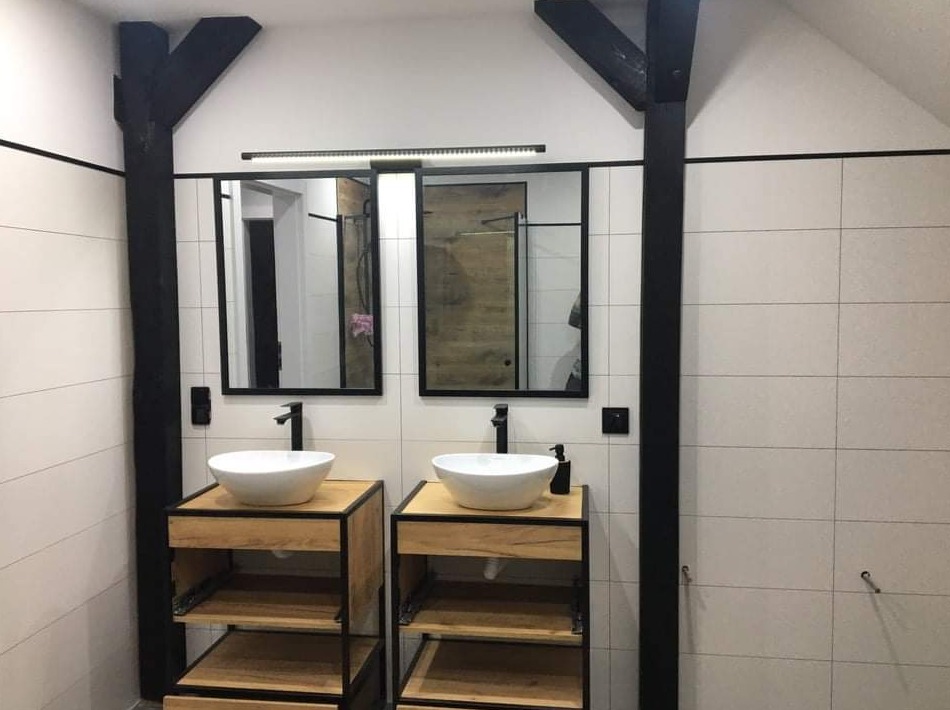 Nowoczesna łazienka z dwoma umywalkami nablatowymi na drewnianych szafkach z czarnymi ramami, czarne ramy luster i belki sufitowe, białe płytki na ścianach.