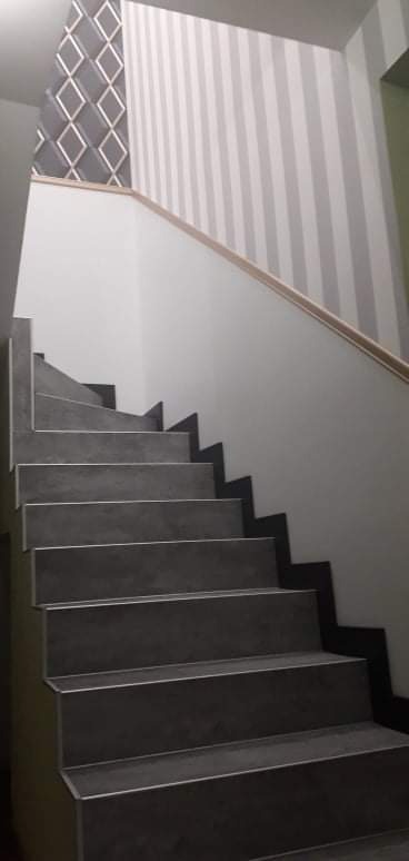 Schody z szarych płytek, wykończone listwą, na ścianie tapeta w szaro-białe pasy oraz wzór geometryczny w romby.