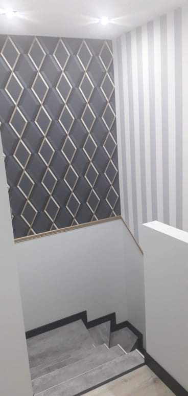 Klatka schodowa z szarą tapetą w romby z metalicznymi obwódkami po lewej i tapetą w szaro-białe pionowe pasy po prawej stronie, szare schody z czarnymi wykończeniami.