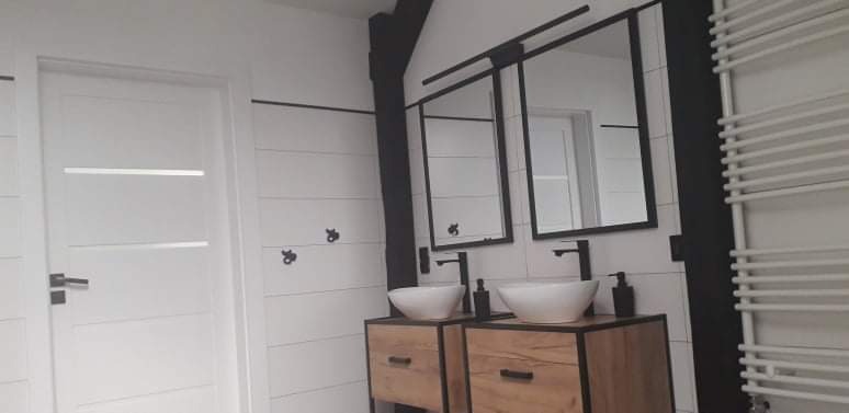 Nowoczesna łazienka z drewnianymi szafkami pod umywalki w kształcie mis, czarne ramy luster i armatura, białe ściany z poziomymi panelami i dekoracyjnymi haczykami, biały grzejnik drabinkowy.