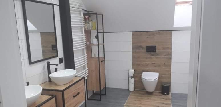 Nowoczesna łazienka z podwójną umywalką nablatową, drewnianymi szafkami z czarnymi ramami, grzejnikiem drabinkowym i toaletą na tle drewnopodobnych płytek.