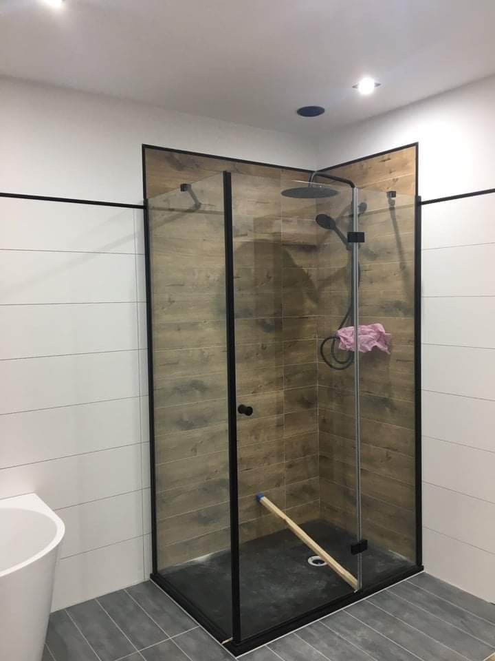 Nowoczesna kabina prysznicowa z czarnymi profilami i panelami imitującymi drewno, z widoczną deską opartą o ścianę, sugerującą prace wykończeniowe.