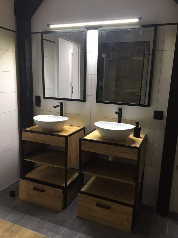 Dwie umywalki nablatowe w stylu loft, osadzone na drewnianych szafkach z czarnymi metalowymi ramami, podświetlane lustra w czarnych ramach, szare płytki podłogowe.