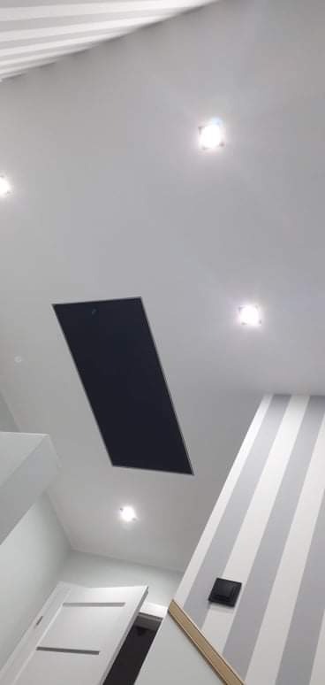 Biały sufit z czarnym panelem rewizyjnym, oświetleniem punktowym LED i fragmentem ściany w biało-szare pasy w nowoczesnym wnętrzu.