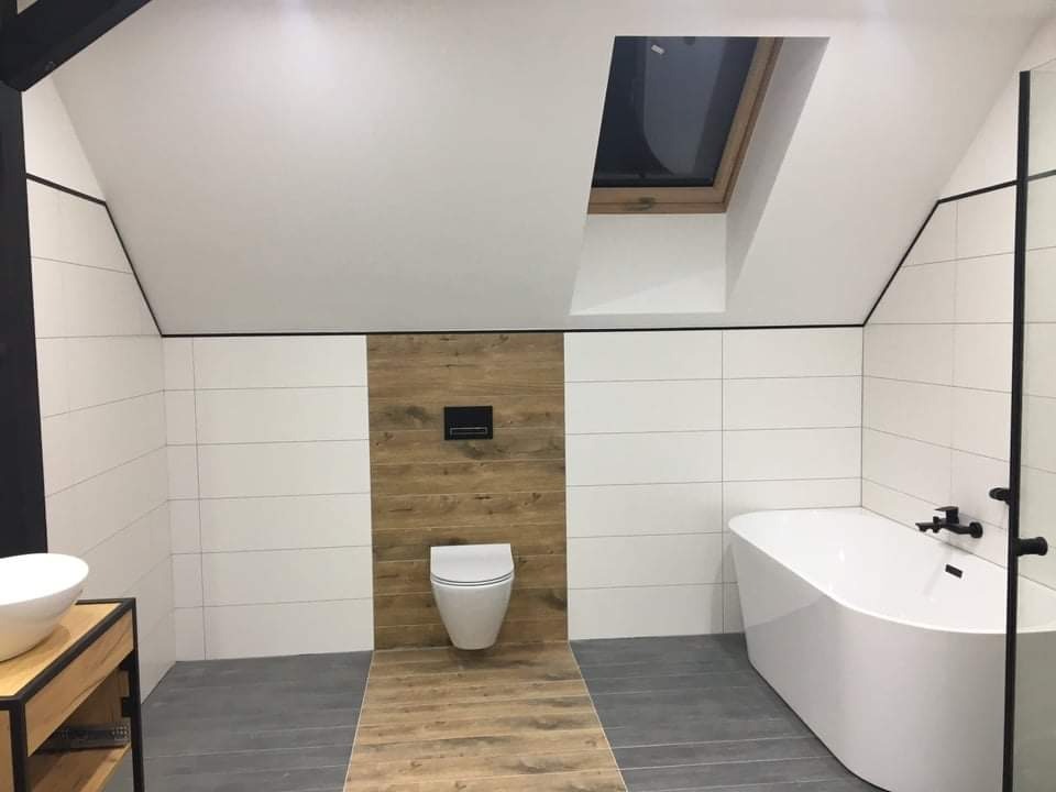 Nowoczesna łazienka na poddaszu z drewnianym akcentem, białą armaturą i płytkami imitującymi drewno, oświetlona przez okno dachowe.