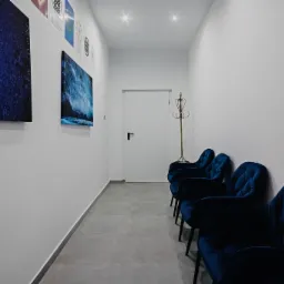 Jasny korytarz poczekalni z błękitnymi fotelami, na ścianach wiszą obrazy o tematyce kosmicznej i morskiej, minimalistyczny wieszak na ubrania przy białych drzwiach.