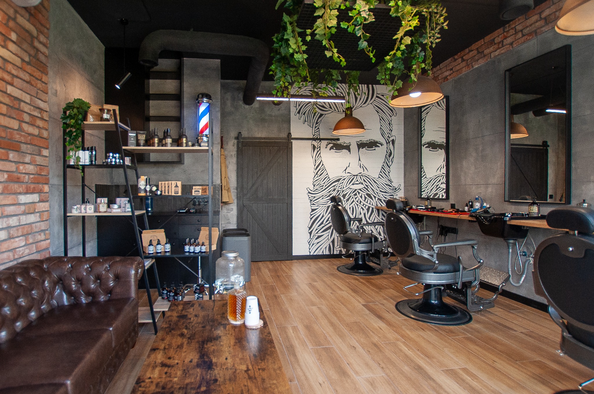 Wnętrze nowoczesnego salonu barberskiego z elementami loftu: ceglana ściana, skórzana sofa, drewniany blat, stanowiska fryzjerskie z lustrami, dekoracja ścienna z wizerunkiem brodacza, wiszące...