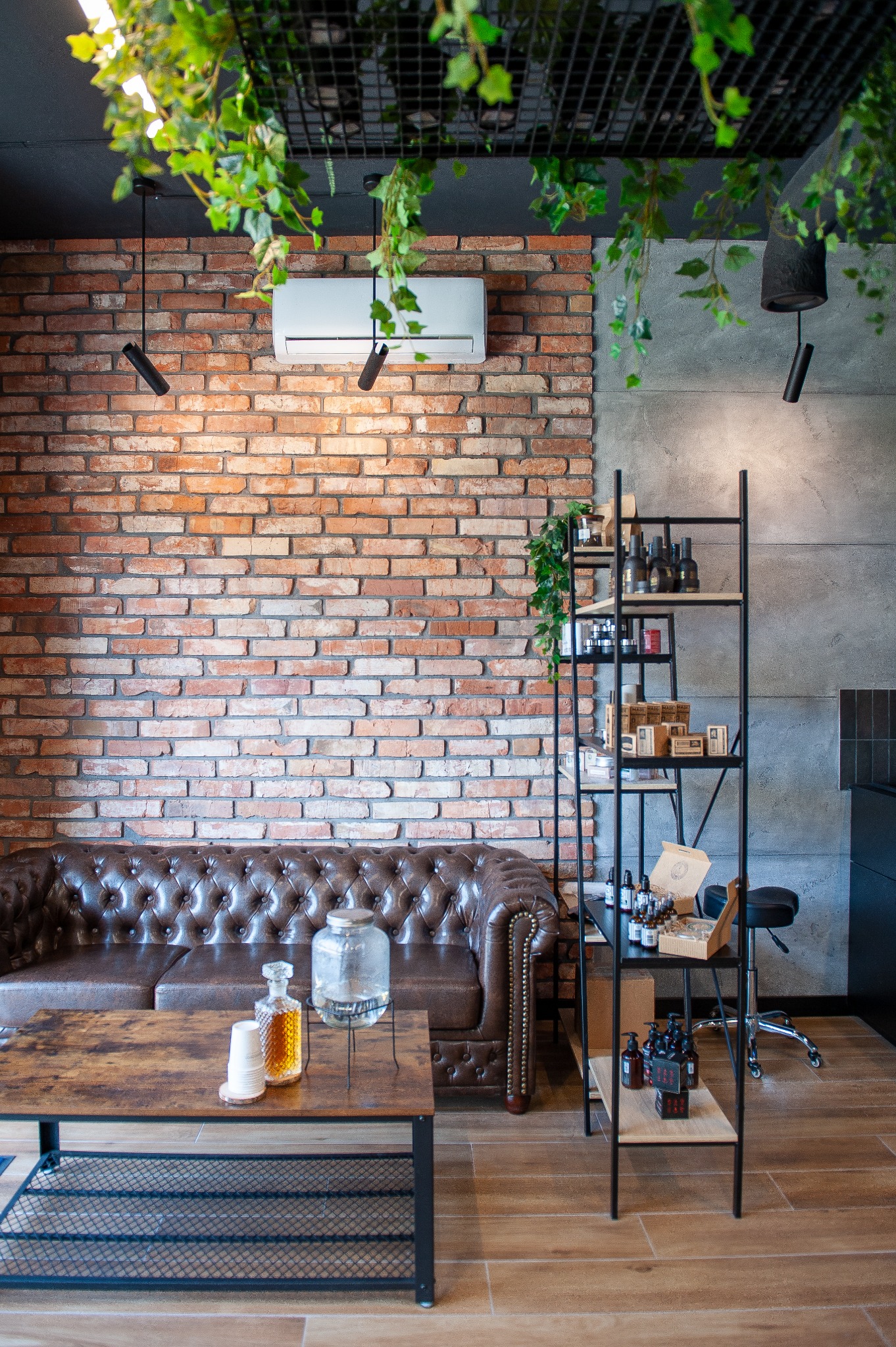 Część poczekalni: skórzana sofa Chesterfield, stolik kawowy z napojami, metalowy regał z kosmetykami, ceglana ściana z klimatyzatorem i wiszącymi roślinami.