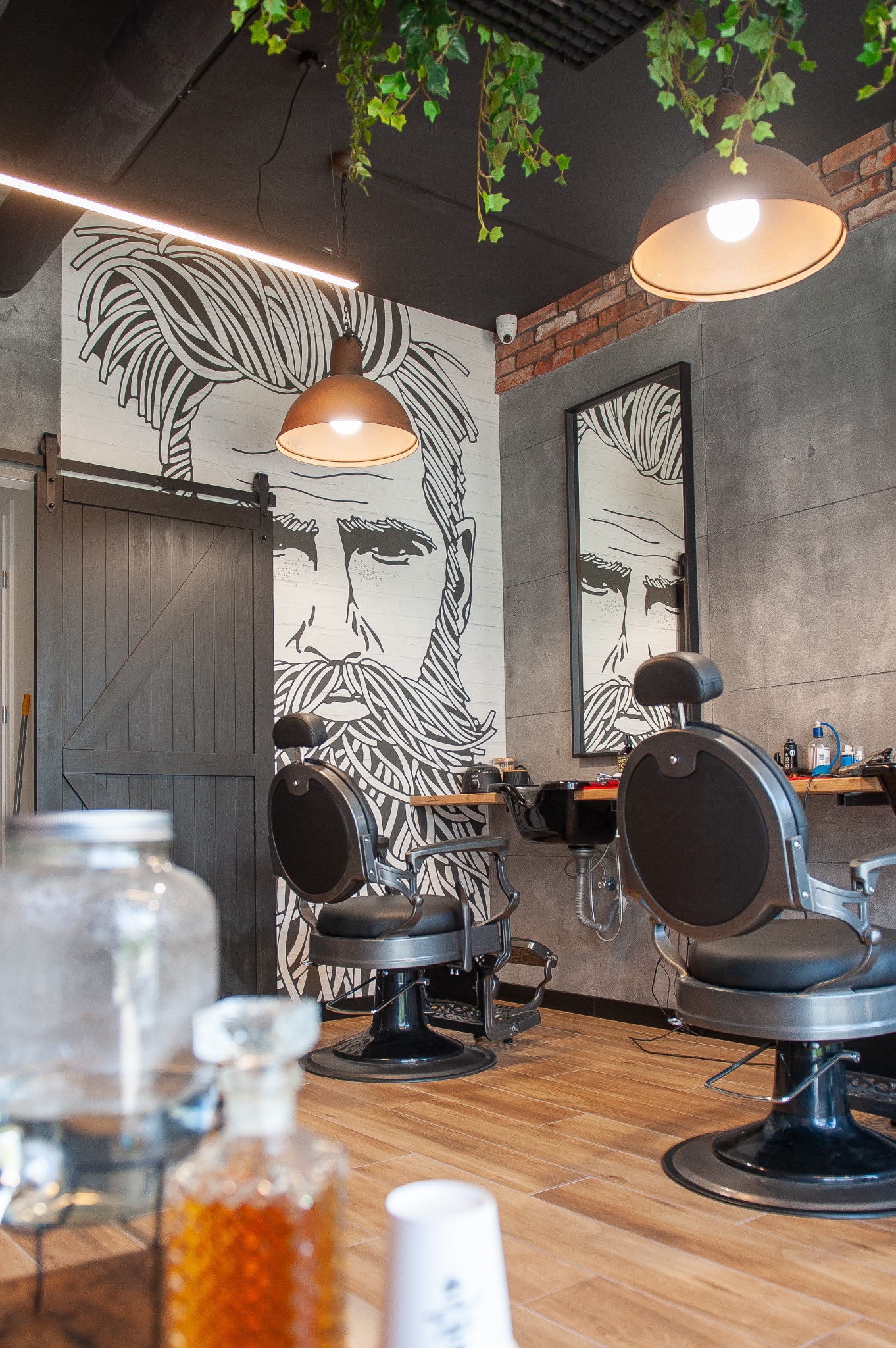 Nowoczesne wnętrze salonu fryzjerskiego z dwoma fotelami, dużym muralem przedstawiającym twarz mężczyzny z brodą, oraz elementami dekoracyjnymi z roślinności.