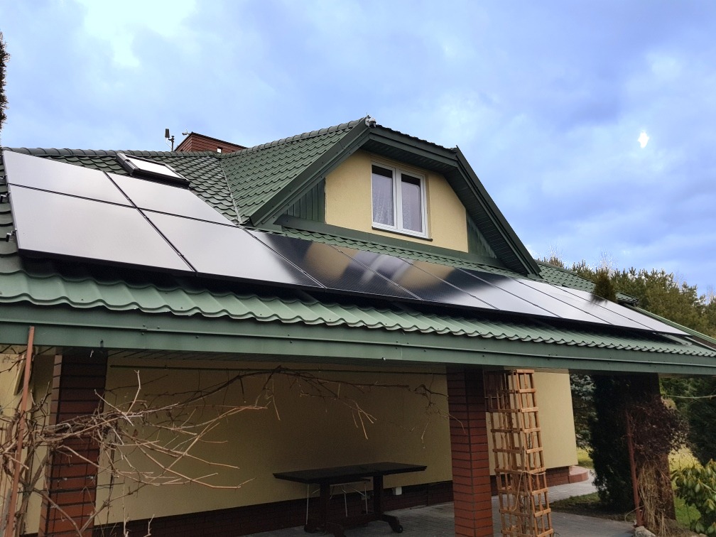 Panele fotowoltaiczne na dachu domu z zieloną blachodachówką, widoczne okno dachowe i fragment ogrodu. Instalacja paneli na dachu skośnym.
