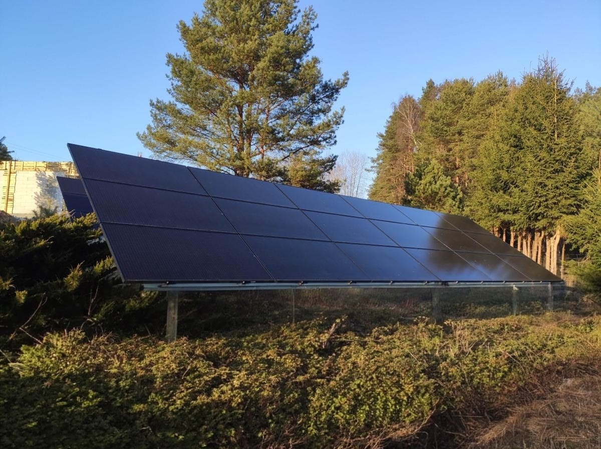 Panel fotowoltaiczny na tle lasu i błękitnego nieba, zamontowany na konstrukcji naziemnej, widoczny w otoczeniu zielonej roślinności.